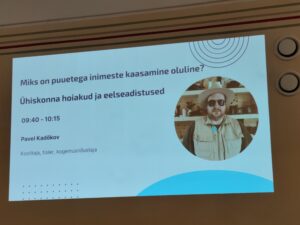 Fotol on pilt seinal olevast slaidist, kus on pealkiri "Miks on puuetega inimeste kaasamine oluline? Ühiskonna hoiakud ja eelseadistused. Sell all esitluse kellaaeg ja esitleja nimi "Pavel Kadõkov". Paremal pool on ringi sees tema foto - mustade päikeseprillidega mees, selgas beež vest ning peas beež kaabu.