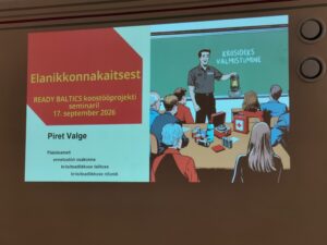 Fotol on pilt seinal olevast slaidist, kus on pealkiri "Elanikonnakaitsest". Selle all tekst "Ready Baltics koostööprojekti seminaril ja kuupäev. Järgmisel real esitleja nimi "Piret Valge Päästeameti ennetustöö osakonna kriisiteadlikkuse talitluse kriisiteadlikkuse nõunik. Paremal pool on illustratsioon, kus Päästeameti töötaja räägib koolitusel osalejatele kriisik valmistumisest. Ümber laua on inimesed ning laua peal erinevad vahendid, priimus, esmaabikarp, konservid.