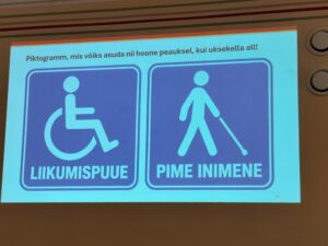Fotol on pilt seinal olevast slaidist, kus on piktogrammid, mis võiksid olla nii hoone peauksel kui uksekella all. Vasakpoolsel piktogrammil on sinise valgega kujutatud ratastoolis inimest ning all on tekst "LIIKUMISPUUE" . Parempoolsel piktogrammil on sinise valgega kujutatud valge kepiga inimest ning all on tekst "PIME INIMENE" .